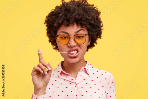 Fotografie Brutal attractive dark skinned female with Afro hairstyle, stretches chewing gum and looks with displeased expression, wears trendy sunglasses, isolated over yellow background