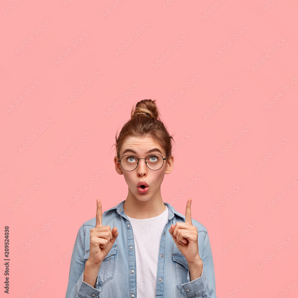 Amazed European female looks upwards with wonderment, indicates with both fingers up, has surprised expression, dressed in fashionable clothing, isolated over pink background with copy space Amazed European female looks upwards with wonderment, indicates with both fingers up, has surprised expression, dressed in fashionable clothing, isolated over pink background with copy space