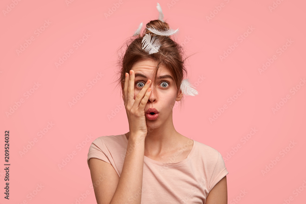 Portrait of suprised cute European young female has no make up, overslept work, woke up late, covers face with hand, peeks through fingers with amazed expression, stands against pink background Portrait of suprised cute European young female has no make up, overslept work, woke up late, covers face with hand, peeks through fingers with amazed expression, stands against pink background