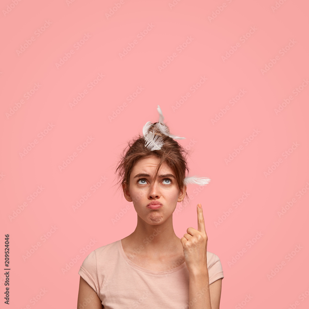 Vertical shot of unhappy beautiful young female with displeased expression, has feather on head, indicates with fore finger upwards, has sleepy look, complains on noisy neighbours who live upstairs Vertical shot of unhappy beautiful young female with displeased expression, has feather on head, indicates with fore finger upwards, has sleepy look, complains on noisy neighbours who live upstairs