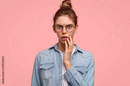 Stupefied beautiful young female with amazed expression and displeased look, keeps hand on face, being puzzled by something unexpected, has attractive appearance, stands against pink studio wall