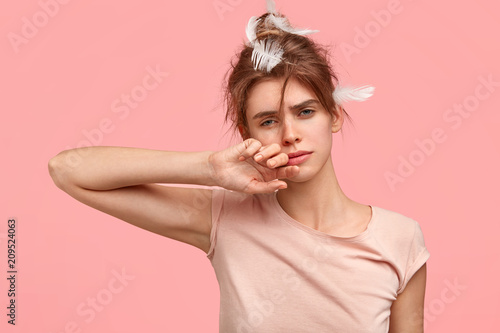 Tired sleepy female rubs eyes and has fatigue expression, wants to have rest, didn`t sleep two nights as worked on diploma paper, has feather on hair, dressed in t shirt in one tone with background photo