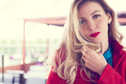 Outdoor fashion portrait of glamour young cheerful stylish lady wearing trendy red coat. Blonde long hair. Style photo with a vintage retro instagram filter