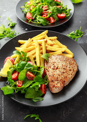 Fried Tuna Steaks on Black Plate with Fresh Green, Tomato Salad, lemon and french fries. healthy sea food