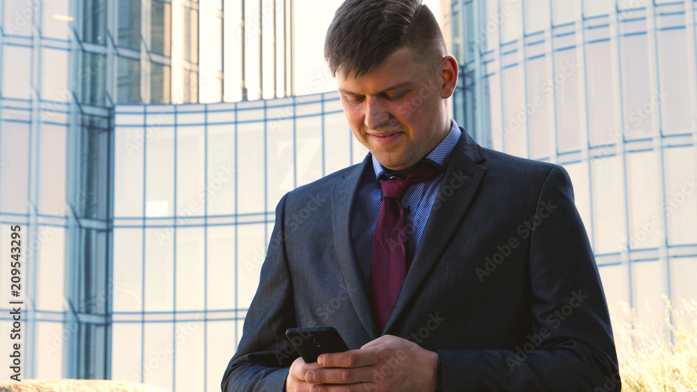 Handsome businessman in suit holding a mobile phone in his hand, writing a message, the background of a skyscraper. Concept: communication, contacts, internet, chat, network. Handsome businessman in suit holding a mobile phone in his hand, writing a message, the background of a skyscraper. Concept: communication, contacts, internet, chat, network.