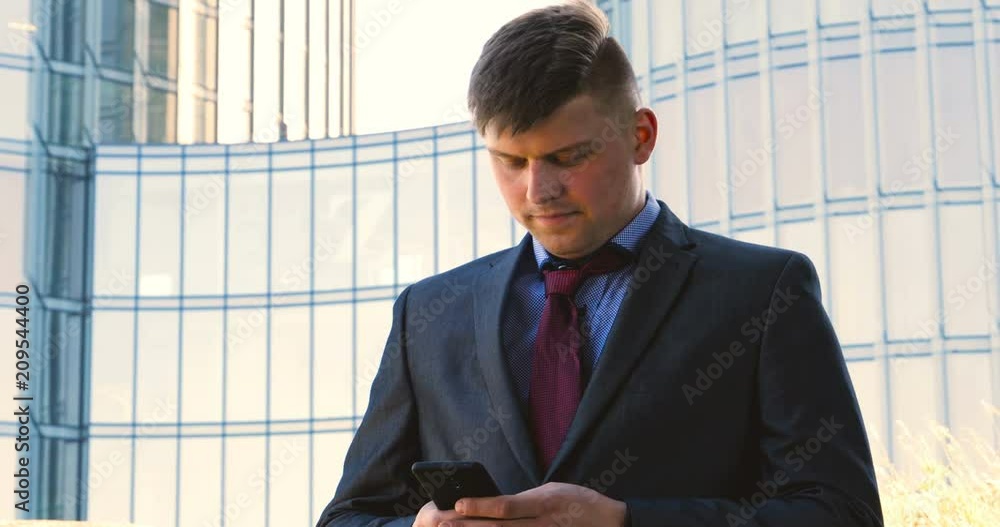 Handsome businessman in suit holding a mobile phone in his hand, writing a message, the background of a skyscraper. Concept: communication, contacts, internet, chat, network. Handsome businessman in suit holding a mobile phone in his hand, writing a message, the background of a skyscraper. Concept: communication, contacts, internet, chat, network.