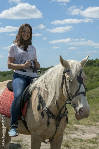 Image of happy female sitting on purebred horse and looking at camera outdoors