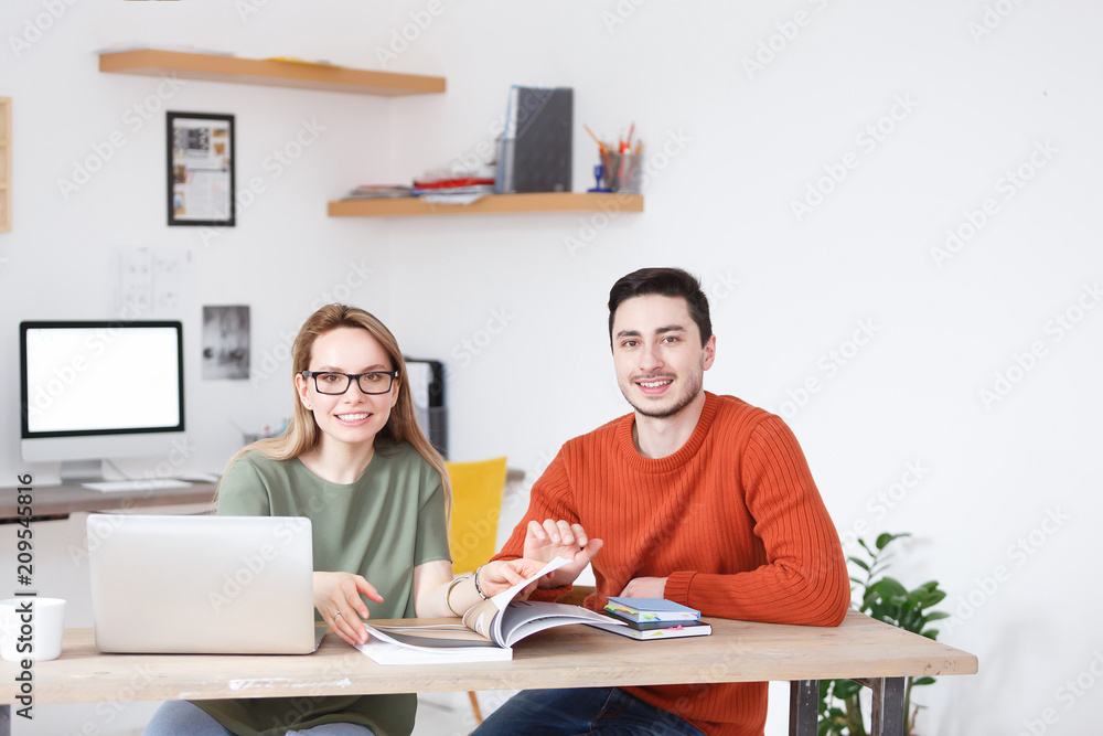 Concert of successful young office workers. Charming cute woman and attractive young male designer or architect sitting at the desk with computers, magazines and notebooks Concert of successful young office workers. Charming cute woman and attractive young male designer or architect sitting at the desk with computers, magazines and notebooks