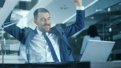 Handsome Businessman Works on a Laptop at His Desk, He Finished Job, Broke the Jackpot, Won Big and Is Happy and Celebrates His Success, Shows His Muscle. Stylish Man in Modern Glass Office. © Gorodenkoff