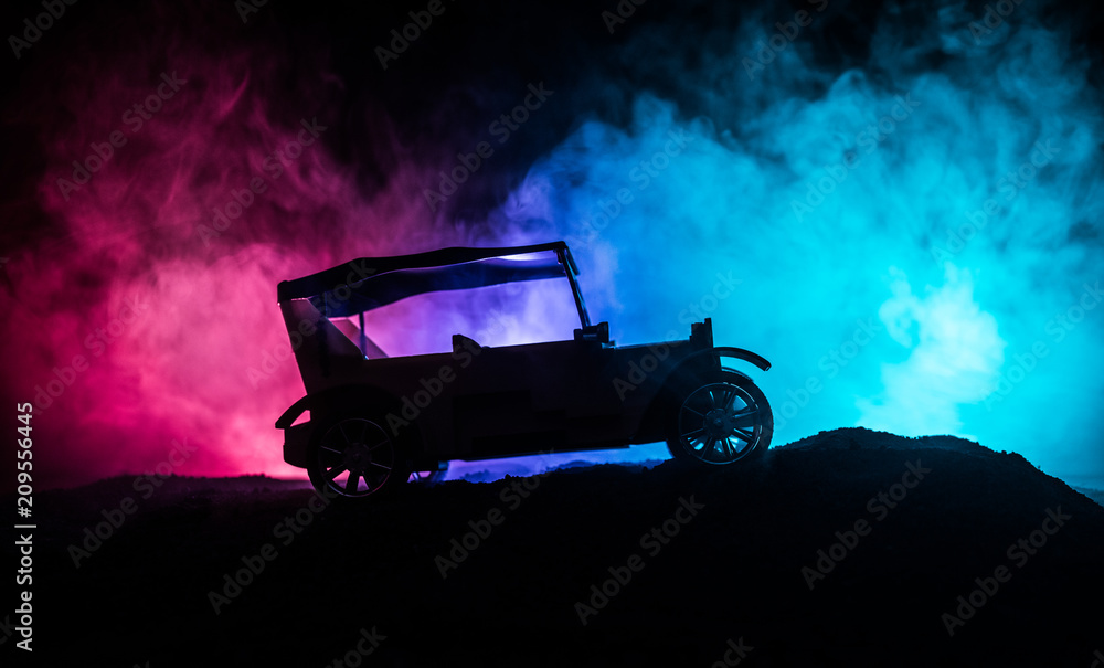 Obraz premium Silhouette of old vintage car in dark foggy toned background with glowing lights in low light.