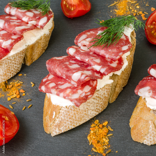 Fresh tasty salami sandwiches. Appetizing sandwiches with smoked sausage, cream cheese and tomatoes. On black wooden background. Square.