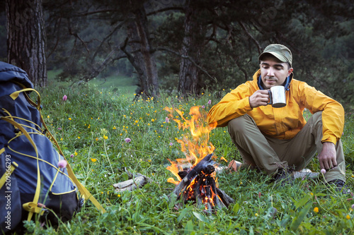 Attractive man drinking hot tea near camp fire at camp in woods