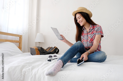 Young asian woman traveler holding map and smartphone while sitting on bed with smiling face, template, travel summer holiday vacation concept
