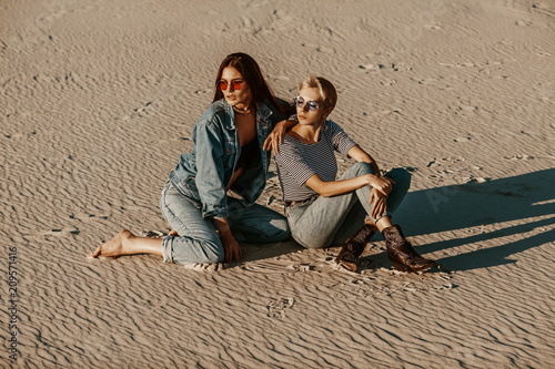 Outdoor portrait of two young beautiful models posing on sand at sunset, wearing stylish denim clothes, round sunglasses. Female fashion concept