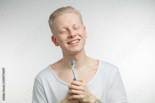 Portrait of smiling young handsome blonde man, floating in clouds with closed eyes, not yet awaken, while keeping toothbrush in tattooed hand. Dental care and teeth health photo