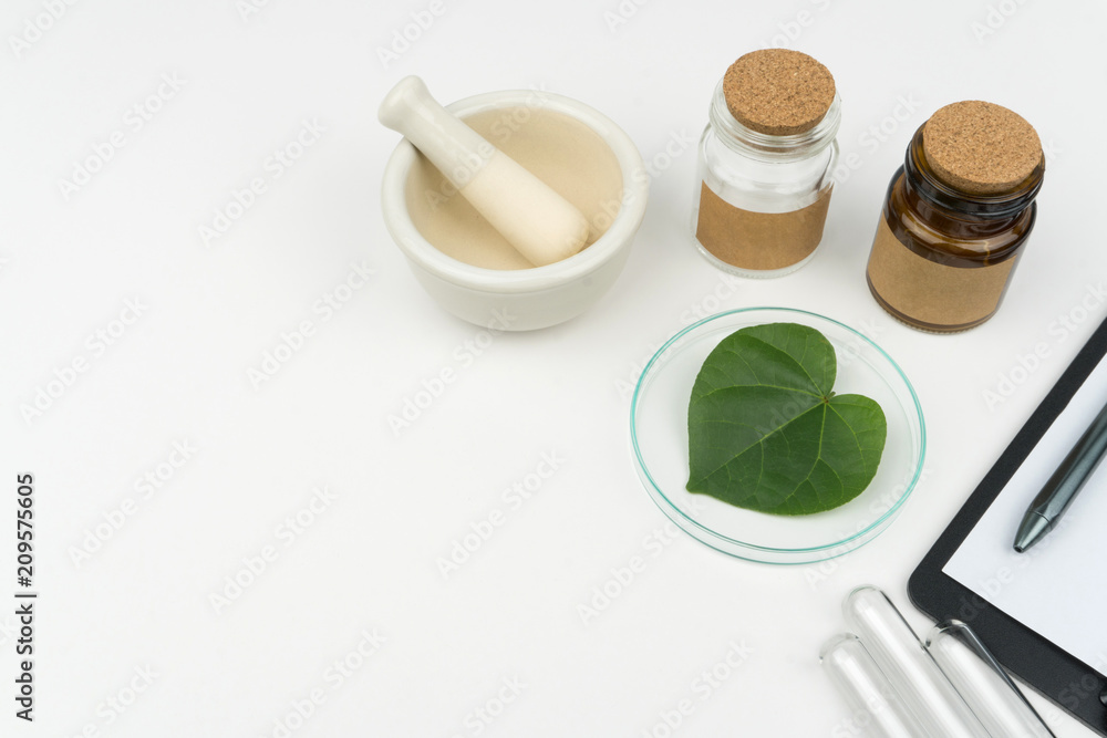 herbal medicine research concept. an organic green leaf in watch glass with a mortar and pestle, two bottles and three test tubes on the white table in laboratory. herbal medicine research concept. an organic green leaf in watch glass with a mortar and pestle, two bottles and three test tubes on the white table in laboratory.