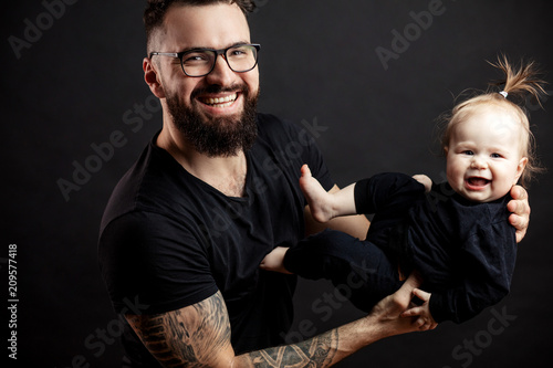 Portrait of happy handsome adult father in perfect fit, wearing spactacles, holding his cute laughing infant daughter in hands, looking at camera, over black background. photo