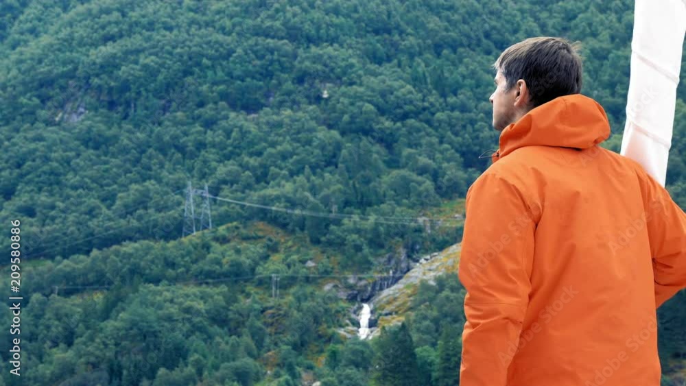 Back view from the side Low Angle. Man in warm jacket looks at high beautiful mountain, standing on white yacht during a cruise. Wonderful landscape of the Norwegian Fjord. Power lines on the mountain Back view from the side Low Angle. Man in warm jacket looks at high beautiful mountain, standing on white yacht during a cruise. Wonderful landscape of the Norwegian Fjord. Power lines on the mountain