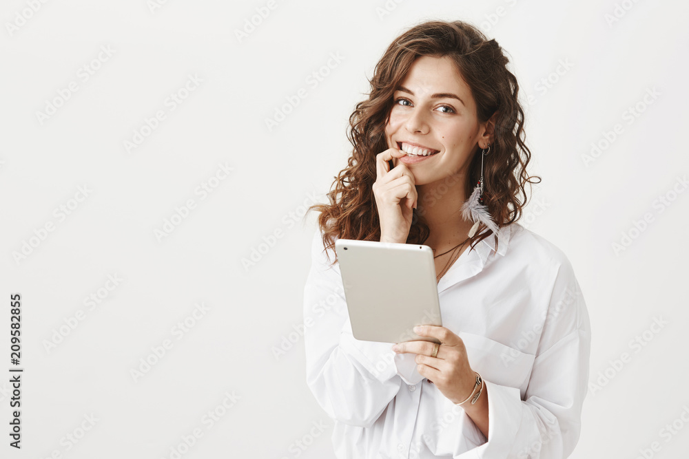 Portrait of charming woman with curly hair biting finger and smiling sensually at camera while holding tablet and standing over gray background. Boyfriend making breakfast while girl plays in gadget Portrait of charming woman with curly hair biting finger and smiling sensually at camera while holding tablet and standing over gray background. Boyfriend making breakfast while girl plays in gadget