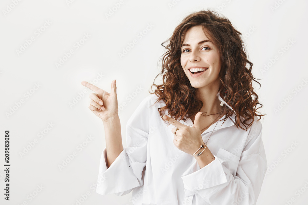 Portrait of happy successful caucasian female with curly hair pointing left with both hands, smiling at camera, standing against gray background. Woman uses all her charm to invite guy for a drink Portrait of happy successful caucasian female with curly hair pointing left with both hands, smiling at camera, standing against gray background. Woman uses all her charm to invite guy for a drink