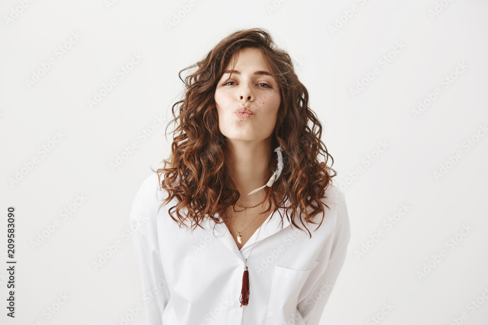 Charming cute caucasian woman with trendy haircut and feather earring bending towards camera to give a kiss, standing against gray background. Attractive girlfriend send off guy to work Charming cute caucasian woman with trendy haircut and feather earring bending towards camera to give a kiss, standing against gray background. Attractive girlfriend send off guy to work