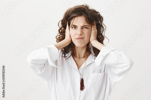 Attractive adult woman covering ears with hands and expressing annoyens and dislike, squinting from awful sound, standing over gray background. Girl hates when someone cracks joints of fingers