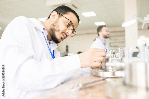 Profile view of concentrated Arabian engineer wearing lab coat sitting at desk and assembling pressure sensor  interior of measuring device factory on background