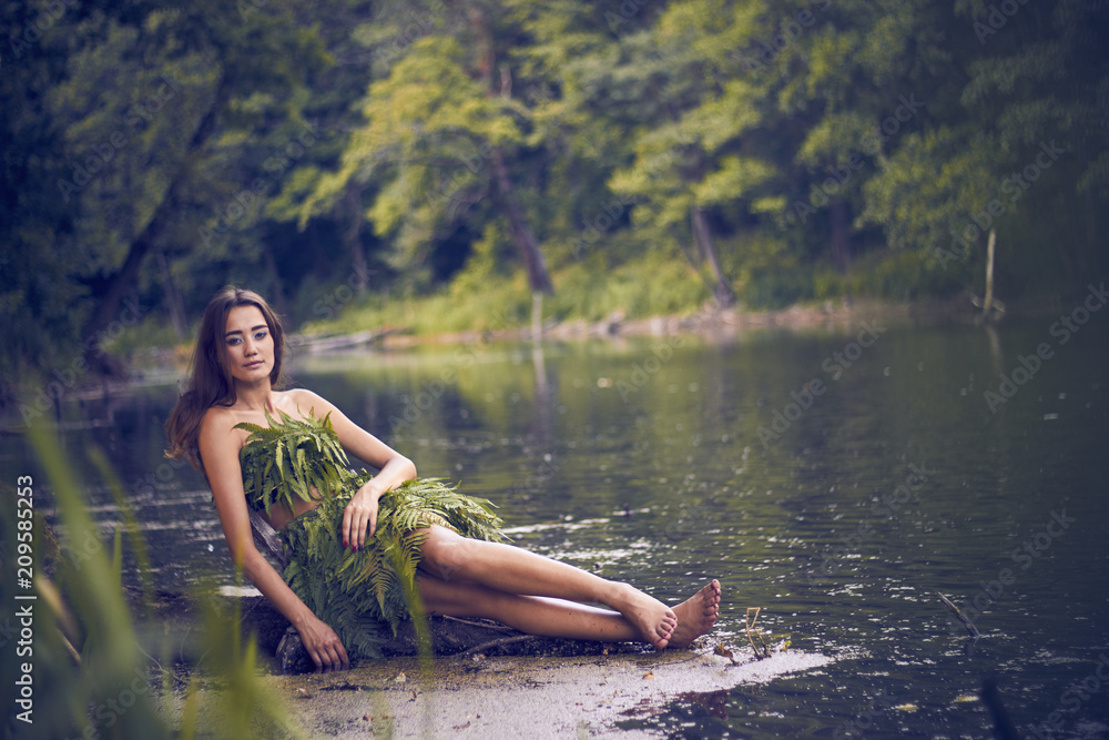 Fototapeta premium Summer heat. Beautiful young girl in a forest lake, open clothes from fern leaves, perfectly combined with nature. A person in the form of a fantasy elf, dryads, cosplay of a character.