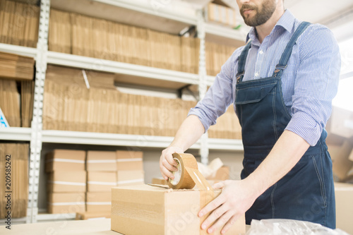 Bearded worker wearing denim jumpsuit using adhesive tape while packing production unit in cardboard box, interior of factory warehouse on background photo