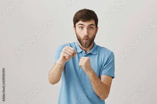 Punch from right and you are in knock out. Studio shot of funny emotive caucasian male with beard raising fists and pulling hand at camera as if hitting it, standing over gray wall in playful mood