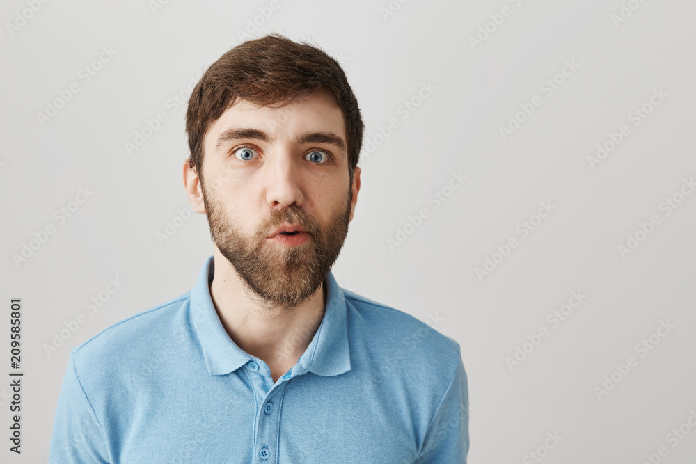 You must be joking. Portrait of confused cute bearded guy, standing with doubtful and questioned expression, moving jaw and looking with popped eyes at camera while posing over gray background