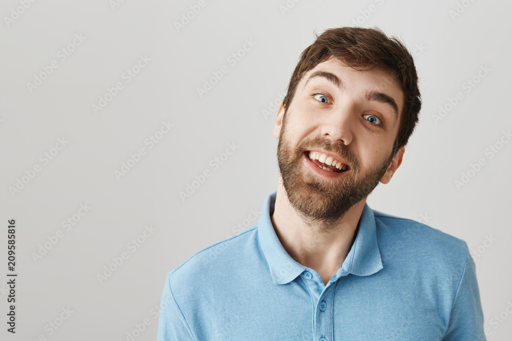 Close-up shot of cute positive european male with beard and moustache, smiling joyfully and staring with crazy look at camera, standing over gray background. Brother fools around with baby sister