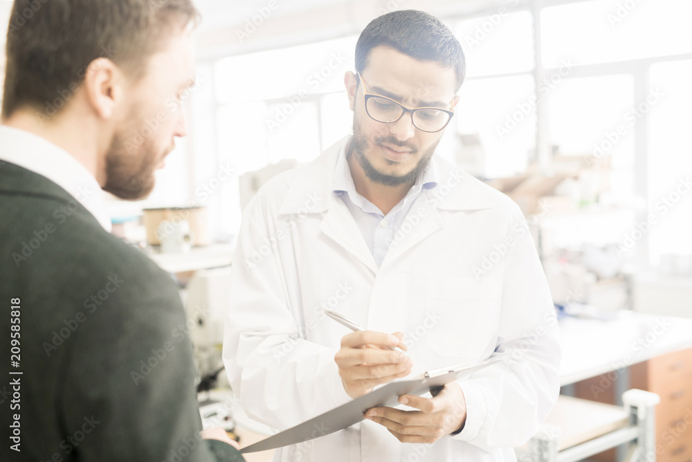 Young Arabian inspector taking necessary notes while bearded entrepreneur giving tour of modern factory, blurred background