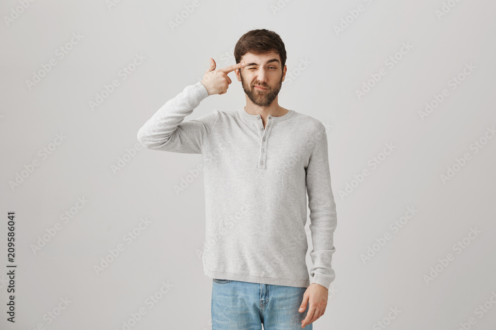 Tired or bored good-looking guy holding gun gesture near temples and making faces while standing over gray background. Upset man can not handle speaking with his annoying mother-in-law Tired or bored good-looking guy holding gun gesture near temples and making faces while standing over gray background. Upset man can not handle speaking with his annoying mother-in-law