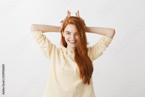 Studio shot of good-looking sassy girlfriend with red hair holding fingers behind head as if ears or devil horns  smiling with interest and curiosity  having idea for night  standing over gray wall