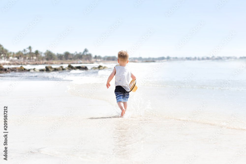 Happy three years old child boy in yellow hat running at white sand and jumping in the waves during summer vacation on tropical beach near blue sea. Smiling and cute toddler kid at the sea Happy three years old child boy in yellow hat running at white sand and jumping in the waves during summer vacation on tropical beach near blue sea. Smiling and cute toddler kid at the sea