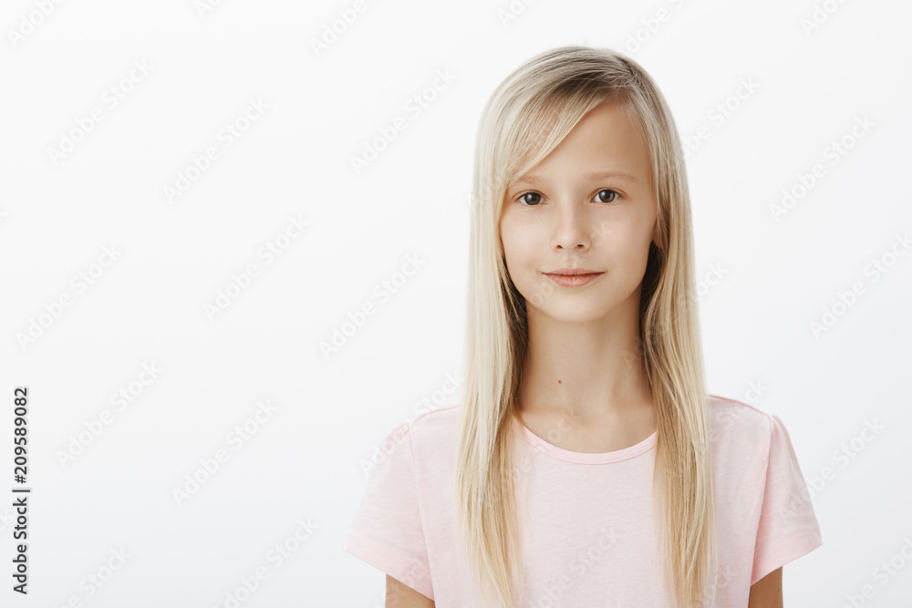Waist-up portrait of relaxed cute female kid with natural blond hair in pink t-shirt, standing confident and relaxed over gray background, smiling slightly, being in good mood, waiting in queue Waist-up portrait of relaxed cute female kid with natural blond hair in pink t-shirt, standing confident and relaxed over gray background, smiling slightly, being in good mood, waiting in queue