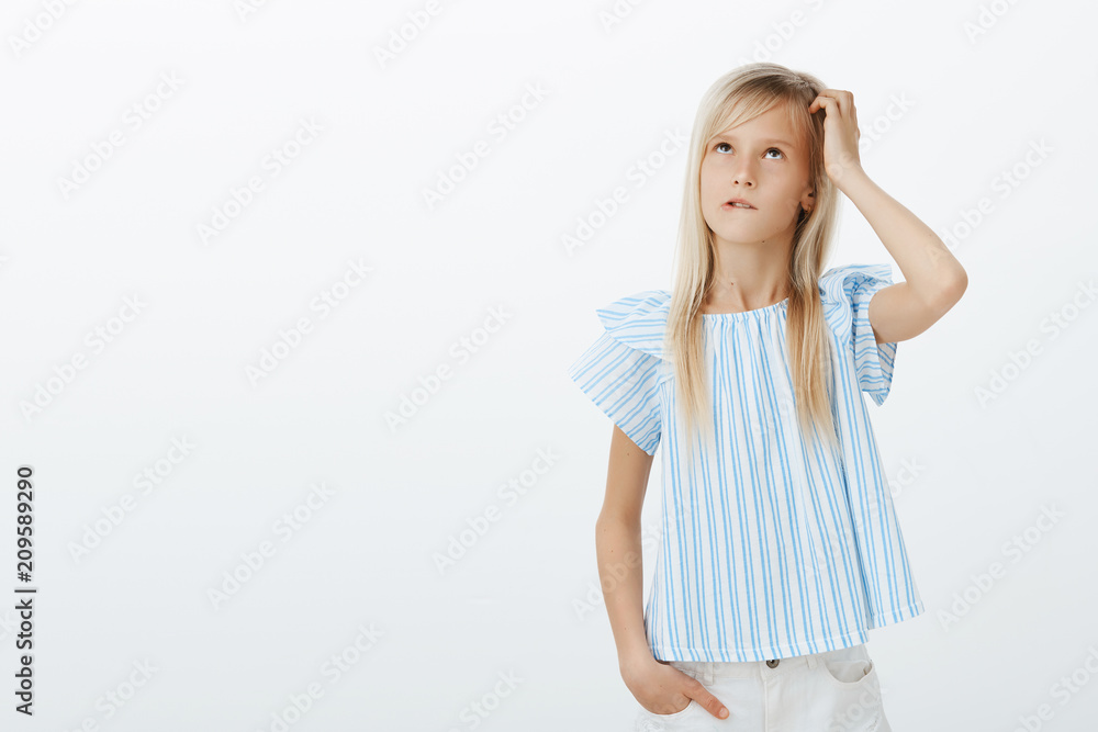 Focused clueless girl trying to calculate in mind near blackboard. Portrait of confused questioned kid in stylish blue blouse, scratching head and looking up, biting lip while thinking over gray wall Focused clueless girl trying to calculate in mind near blackboard. Portrait of confused questioned kid in stylish blue blouse, scratching head and looking up, biting lip while thinking over gray wall
