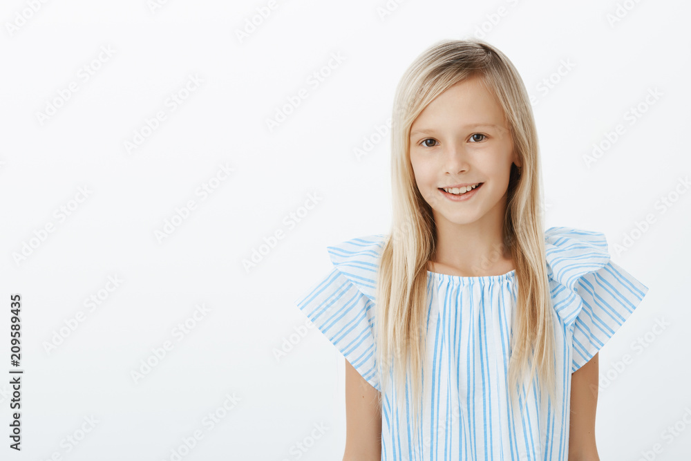 Charming happy young girl introducing herself to new classmates. Portrait of pleased carefree adorable daughter with blond hair in blue blouse, standing casually over gray wall and smiling friendly Charming happy young girl introducing herself to new classmates. Portrait of pleased carefree adorable daughter with blond hair in blue blouse, standing casually over gray wall and smiling friendly