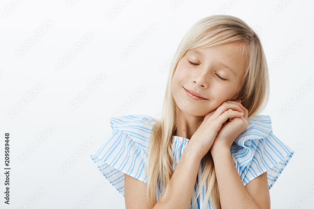 Fototapeta premium Time to sleep. Sleepy cute little european girl with blond hair, closing eyes and leaning on palms as if sleeping, feeling pleased and tired after spending awesome time with friends over gray wall