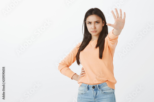 Displeased calm and confident female student in casual clothes, holding hand on hip and pulling palm towards camera in no, stop or enough gesture with strict serious expression over gray wall