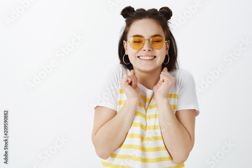 Excited very happy woman in fashionable outfit and glasses, closing eyes and grinning from happiness, clenching fists near chest and smiling broadly, being amazed while waiting for great things happen