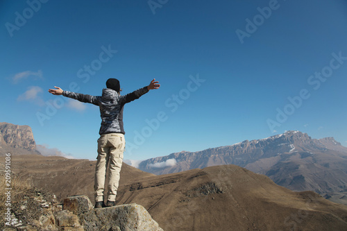 an applause of a young woman backpacker on top of a sprawling upward mountain peak. Freedom and victory against the background of mountains and blue sky