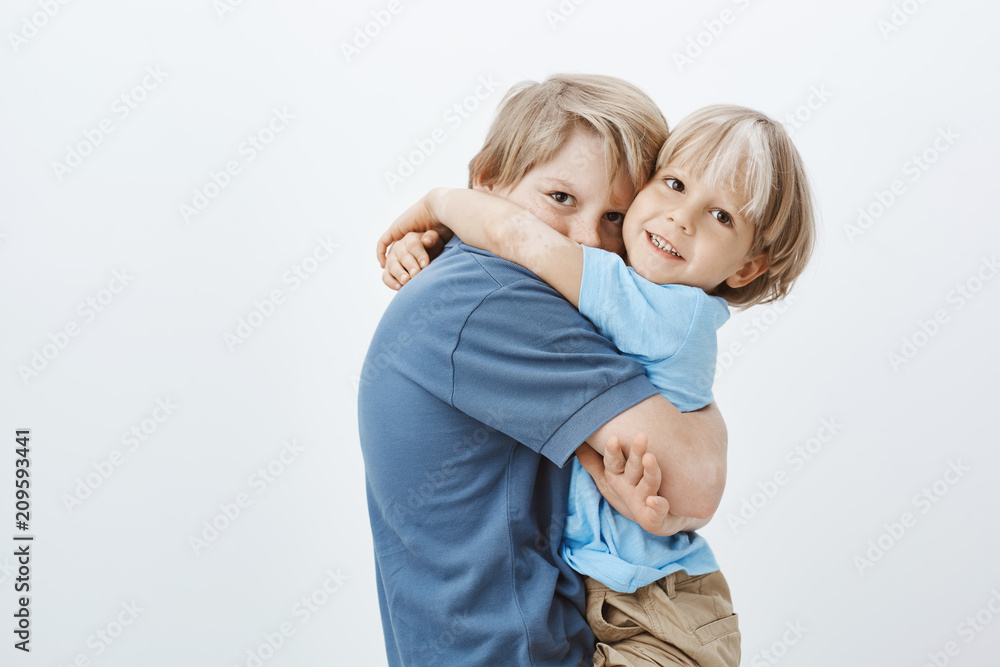 Brothers are best friends. Portrait of cute european boy hugging brother and gazing at camera, being happy to have sibling, loving spending time in family circle and standing over grey wall