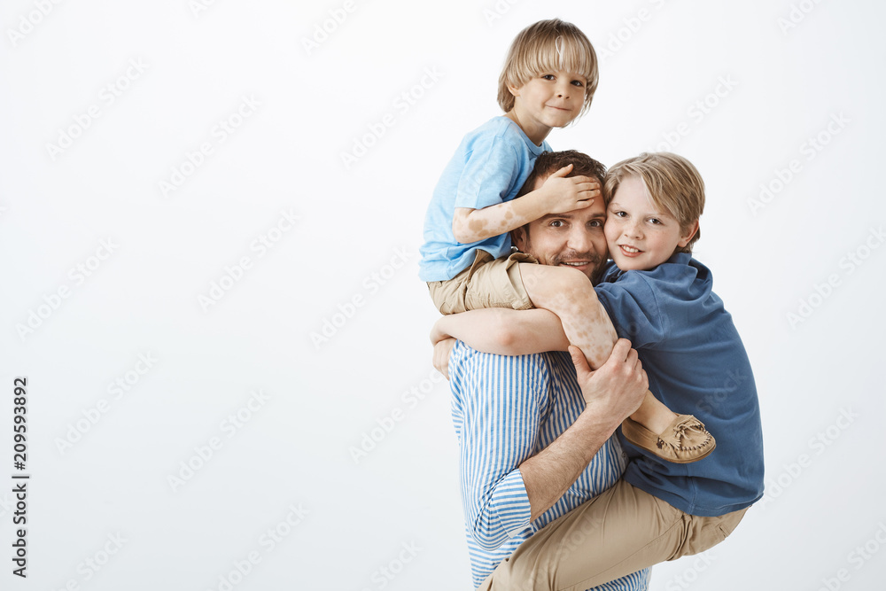 Indoor shot of positive happy family guy holding son with vitiligo on shoulders and cute kid on chest, smiling broadly, feeling joyful while playing with loving children, looking at mom with care