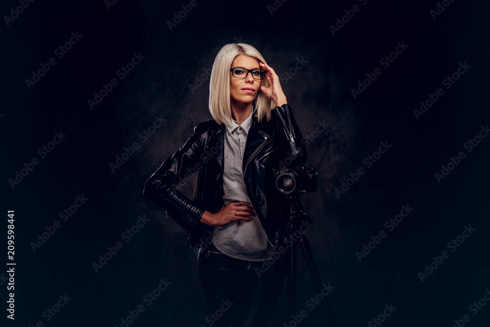 Smart blonde female photographer in trendy clothes posing while leaning on a professional camera with a tripod at a studio, looks at the camera. Smart blonde female photographer in trendy clothes posing while leaning on a professional camera with a tripod at a studio, looks at the camera.