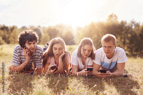 Horizontal shot of four friends of same age, focused into screens of mobile phones, have different facial expressions, lie on stomaches, enjoys summer sunny day and recreation time. Technology concept © sementsova321