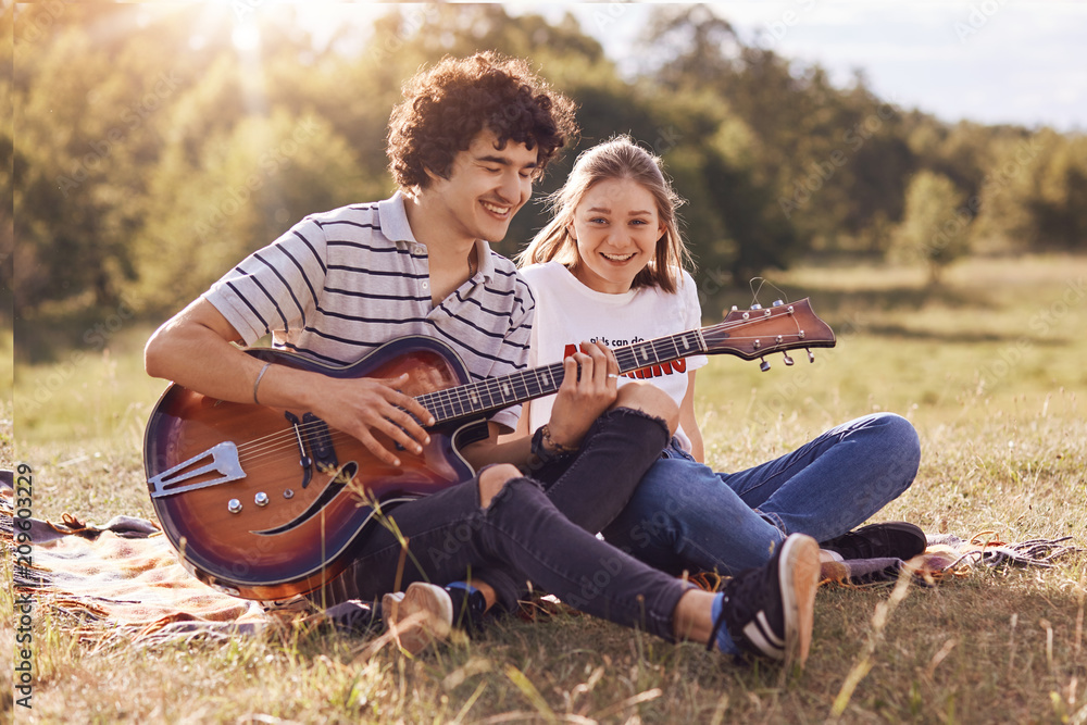 Youth, dating and recreation time concept. Happy girlfriend and boyfriend sit on green grass, play guitar, enjoy calm atmosphere, have smiles on faces. Lovely young female listens romantic songs