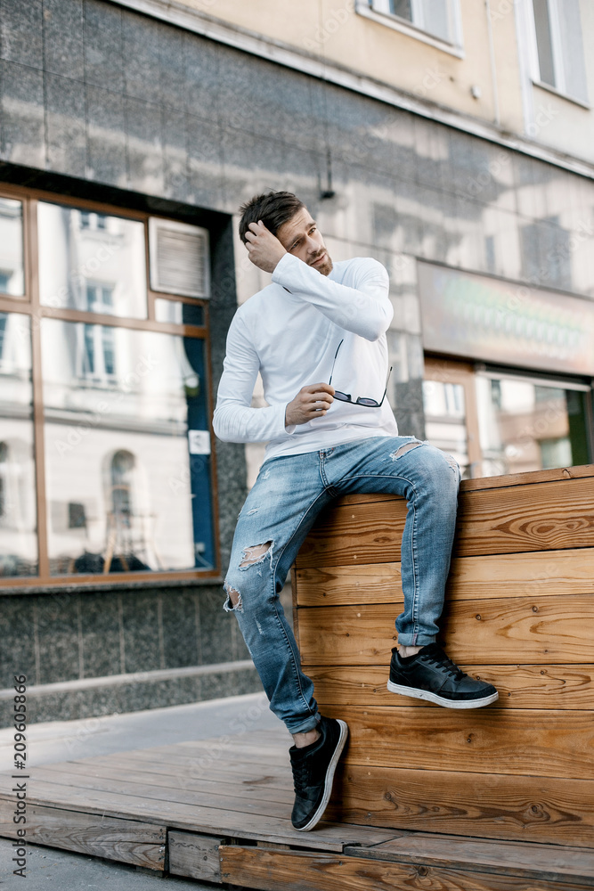 A young man travels through the streets of the city. Boy in sunglasses Stylish man near the building. The man sits on a whip. Beard and hair in a stylish guy.