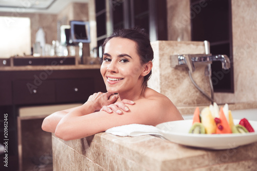 Happy young female lying in bath with folded hands. She is leaning on towel with smile and having fruit plate beside her 